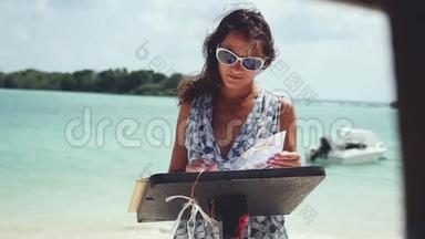 年轻的黑发女人戴着太阳镜在海滩餐厅看菜单。 她身后有船的大海景色